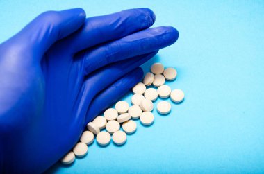 Mavi eldivenli bir erkek, makro mavi arka planda beyaz tabletlerle tırmıklıyor. El ve haplar