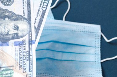 Bir tıbbi mavi maske siyah arka planda 100 dolar değerinde dolarların üzerinde yatar. Maske ve dolar erkeklerin elinde