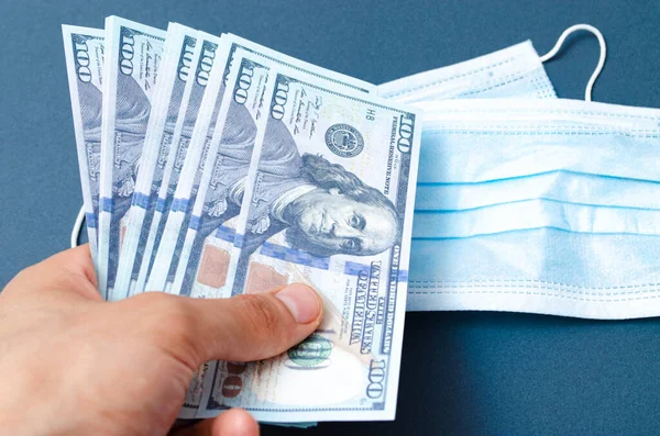 Bir tıbbi mavi maske siyah arka planda 100 dolar değerinde dolarların üzerinde yatar. Maske ve dolar erkeklerin elinde