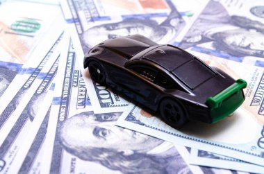 Black toy car on a heap of dollar bills on a white and black background. A sports car with a green spoiler, a wing on dollars in a close-up top and side view. Rear and front car. Selling, buying clipart