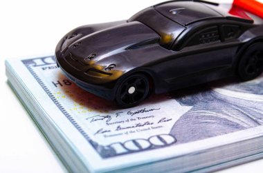 Sıçrama tahtasının üzerinde kırmızı kanadı olan siyah renkli bir spor araba. Dolar banknotlarından yapılmış bir podyum. Araba ve dolar. Piyasada alım, satım fiyatları artıyor