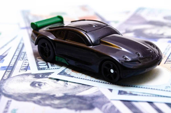 Black toy car on a heap of dollar bills on a white and black background. A sports car with a green spoiler, a wing on dollars in a close-up top and side view. Rear and front car. Selling, buying