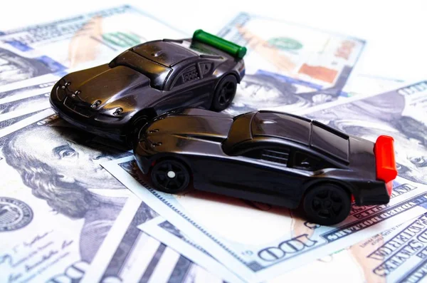 Two black toy cars on a heap of dollar bills on a white and black background. Two sports cars on dollars in close-up. One car overturned, accident, accident. Car insurance