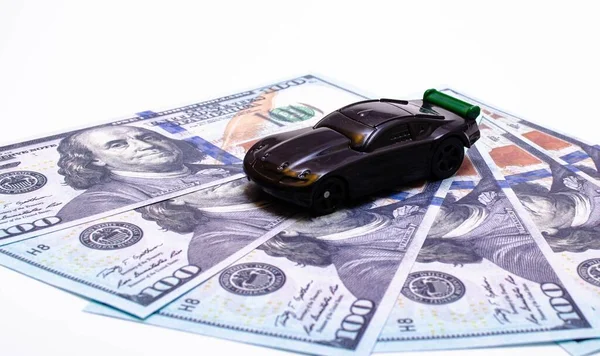 黑色玩具车在一堆美元钞票上的白色和黑色背景 一辆有绿色扰流板的跑车 顶部和侧面都有一个靠美元的机翼 后备箱和前备箱销售 — 图库照片