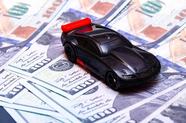 Black toy car on a heap of dollar bills on a white and black background. A sports car with a red spoiler, a wing on dollars in a close-up top and side view. Rear and front car. Selling, buying