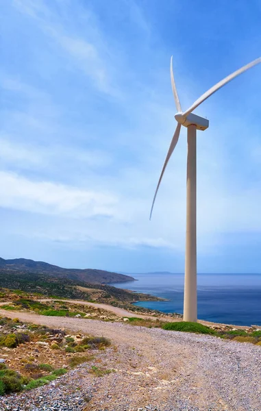 Single windmill turbine on hilltop of seashore in colorful landscape against dynamic blue sky with clouds and winding road.