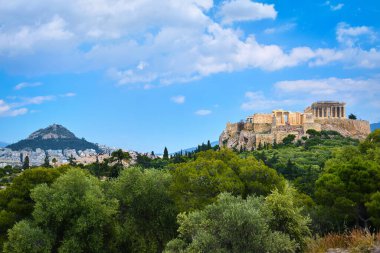 Atina, Yunanistan 'da arka planda Akropolis tepesi ve Lycabettus tepesinin ikonik vew' i yaz güneşinde Pnyx tepesinden mavi gökyüzünde büyük bulutlarla.