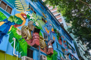 26 Kasım 2019 - Kuala Lumpur, Malezya: Tropikal kuşlar, yapraklar, çiçekler ve salyangozlarla dolu güzel parlak bir duvar resmi. Klima birimleri detaylı sanat eserleriyle harmanlanmış. Güneşli bir günde yapraklı ağaçların altında sokak sanatı.. 