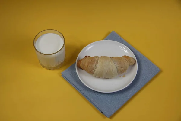 在蓝色餐巾上的盘子里放着羊角面包 在黄色的背景平面上放着一杯牛奶 图像包含复制空间 — 图库照片