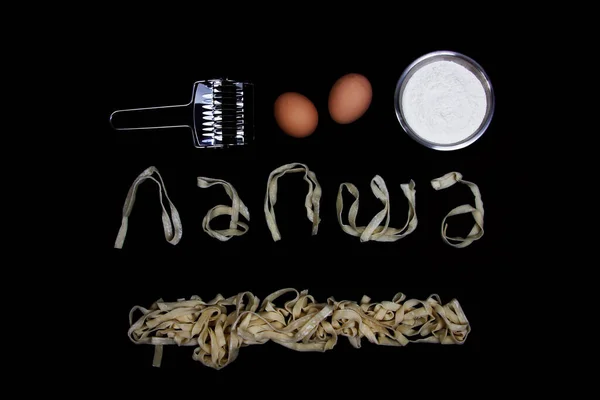 lettering LAPSHA (noodles in russian), made of noodles, eggs, bowl of wheat flour and pastry cutter on black background flat lay. Cooking concept