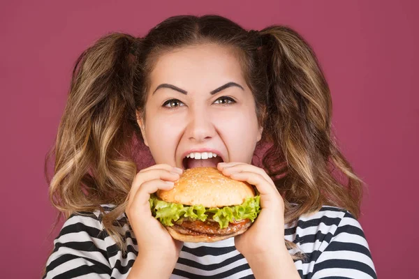 Genç kız mutlu yüz ve Aç açık ağzı ile sığır eti burger sandviç tutarak pembe arka plan izole. Fast food kavramı.