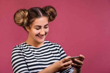 Genç ve güzel hippi kız komik saç modeli ve çizgili gömlek gülümseyen, aygıtı kullanarak akıllı telefon üzerinde pembe arka plan izole holding