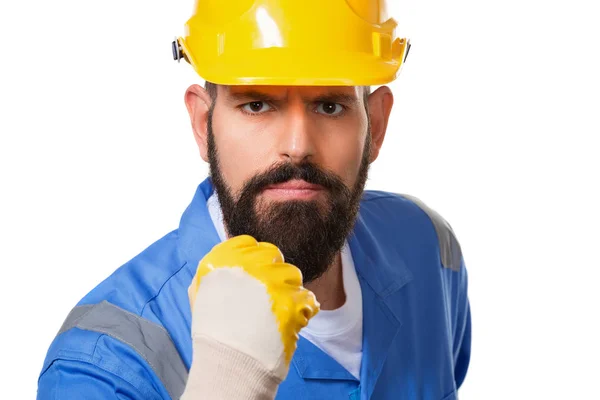 Close up portrait of bearded angry man builder in yellow helmet and blue uniform threatening with fist over white background — Stock Photo, Image