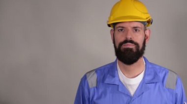 Sarı kask ve mavi üniforma lı sakallı işçi gri arka plan üzerinden kameraya bakıyor