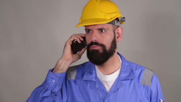 灰色の背景に誰かと電話で話す黄色のヘルメットのひげ労働者 — ストック動画
