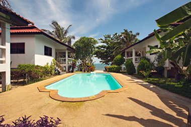 Küçük evler ve turkuaz suyu olan lüks bir havuz, deniz kenarı arka planı.