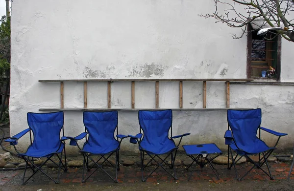 Empty blue folding camping chairs and table lined up in front of a rustic white wall with a ladder on the wall.