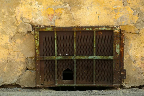 Basement of an old building with falling plaster covered with rusty grid. Street level window block with grid. — Stock Photo, Image