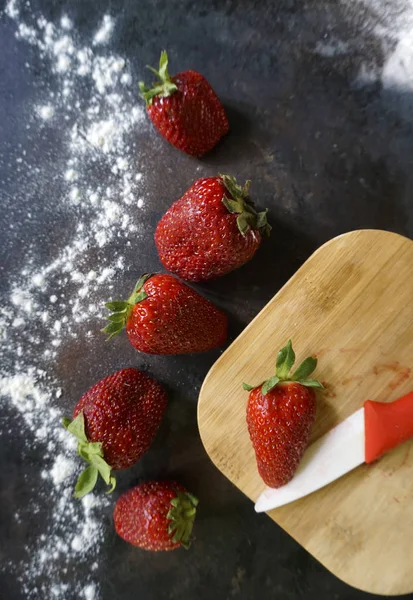 Fraises rouges mûres avec sucre glace en poudre, couteau et planche à découper en bois sur fond sombre. Vue du dessus . — Photo