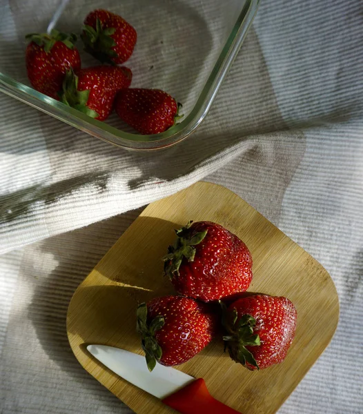 Fresas en tabla de cortar de madera, kinfe y recipiente de vidrio en servilleta de lino blanco . —  Fotos de Stock