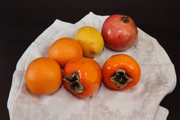 Tropikal meyvelerin hala hayatı. Koyu bir masada yatan bir keten peçete üzerinde Persimmon, narenciye ve nar olgun turuncu meyveler.