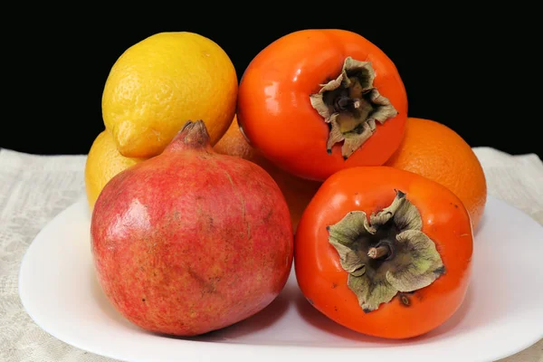 Tropikal meyvelerin hala hayatı. Karanlık bir masada ayakta beyaz servis plaka üzerinde Persimmon, narenciye ve nar olgun turuncu meyveler.