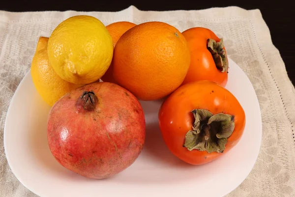 Tropikal meyvelerin hala hayatı. Karanlık bir masada ayakta beyaz servis plaka üzerinde Persimmon, narenciye ve nar olgun turuncu meyveler.