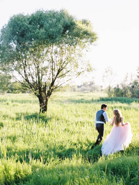 大きな木の近くの緑の芝生で歩く幸せな美しい新婚夫婦の屋外のポートレート — ストック写真