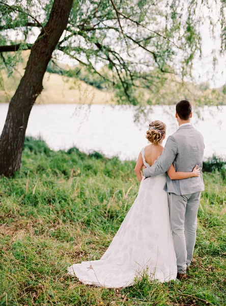 户外肖像快乐美丽的新婚夫妇走在绿色的草坪附近的大树和湖泊 — 图库照片