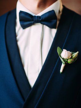 Koyu mavi takım elbiseli şık bir adam, denizci papyonu ve yumuşak gül düğme deliği, damat düğün törenine hazırlanıyor.