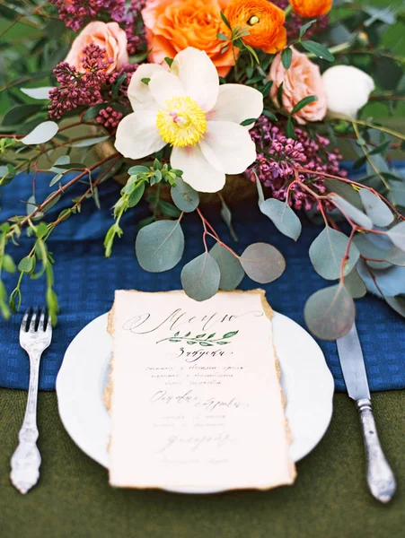 Bright colorful bouquet with exotic flowers on wooden table, menu card on plate and cutlery. Deep blue, orange, yellow, white, green, grey, rosy, purple colors