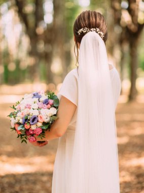 Beyaz elbiseli genç bir gelin ve uzun peçeli yumuşak çiçeklerle düğün buketi tutan yaz ormanı manzaralı.