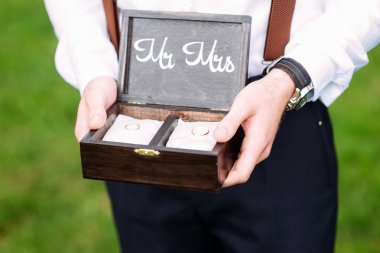 Best man holds wooden box with golden wedding rings on white pillows. Outdoor summer wedding ceremony. Rustic wedding. Mr and Mrs. clipart