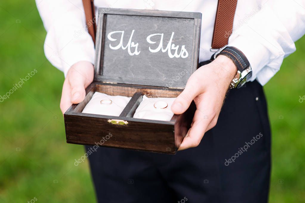 Best man holds wooden box with golden wedding rings on white pillows. Outdoor summer wedding ceremony. Rustic wedding. Mr and Mrs.