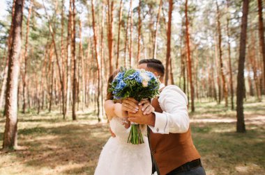 Çam ormanında sonbahar düğünü. Gelin ve damat ellerinde mavi çiçeklerle düğün buketi tutuyorlar. Beyaz, mavi ve kahverengi renkte düğün.