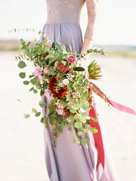 大規模な近代的な紫色のブライダル花束を閉じます 紫のウェディングドレスを着た美しい花嫁が砂浜に立っています バラと赤の花とユーカリの葉で閉じる巨大な結婚式の花束 — ストック写真