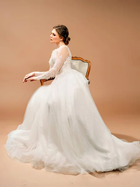 漂亮的新娘穿着白色婚纱坐在一把老式椅子上 带有棕色背景的工作室肖像 时尚婚纱 — 图库照片