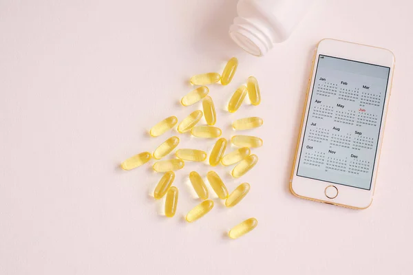 A lot of pills are spilled of a plastic container and a smartphone with calendar screen. Close up, top view with pink neutral background. Vitamin therapy, fish oil, vitamin D.