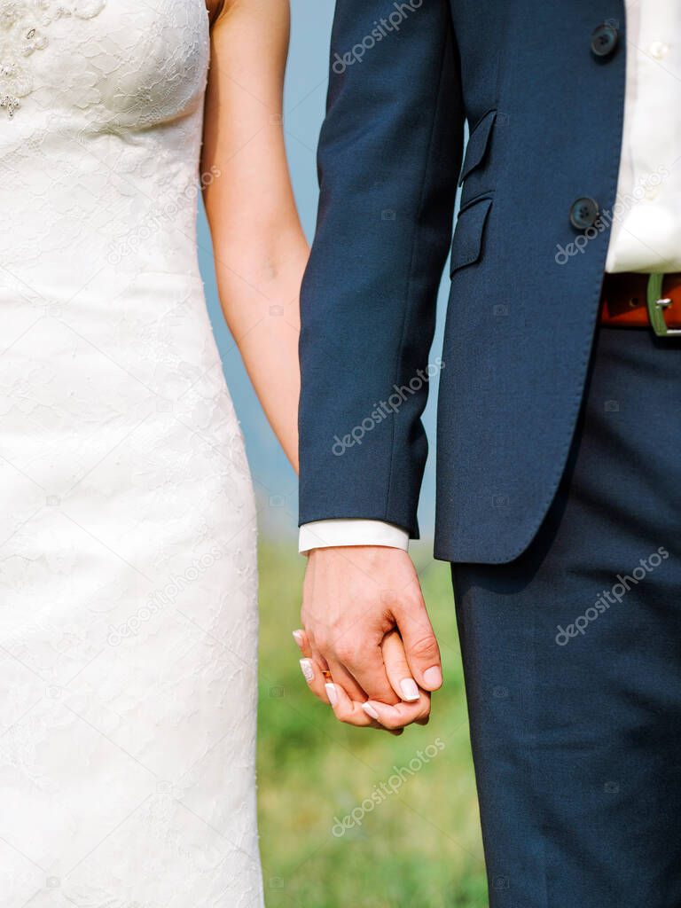 Closeup view of married couple holding hands at outdoor wedding ceremony. Bride in white lace wedding dress and groom in blue suit standing together on green background.