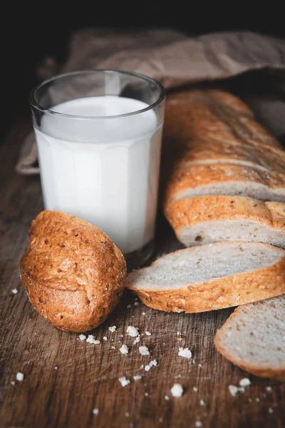 Healthy food. Long loaf of rural bread with two cut-off pieces lie on a wooden chopping board and a glass of fresh milk. Dark background