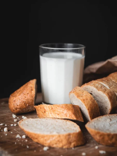 Healthy food. Long loaf of rural bread with two cut-off pieces lie on a wooden chopping board and a glass of fresh milk. Dark background.