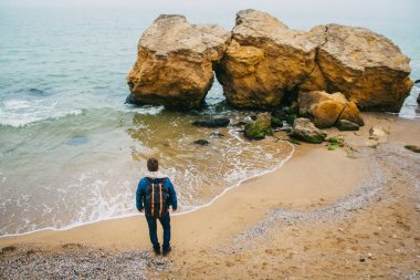 Dalgalar ile güzel bir denize karşı bir kaya nın yanında duran bir sırt çantası ile Gezgin, dünya çapında harika bir yolculuk sırasında sakin bir okyanus yakınında poz şık bir hipster çocuk. Arkadan ateş edin