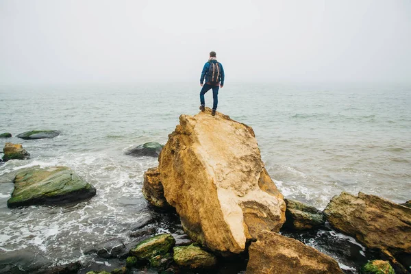 Bir sırt çantası ile Traveler dalgalar ile güzel bir denize karşı bir kaya üzerinde duruyor, şık bir hipster çocuk dünya çapında harika bir yolculuk sırasında sakin bir okyanus yakınında poz. Arkadan ateş edin