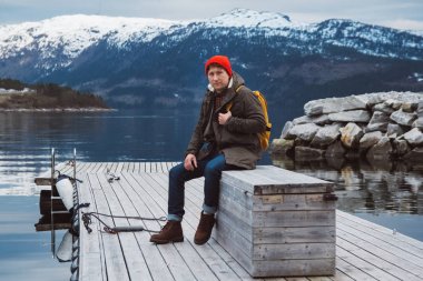 Sarı sırt çantası ile gezgin adam dağ ve göl arka planda ahşap iskele üzerinde oturan kırmızı bir şapka giyiyor. Kısa mesaj veya tanıtım içeriğiniz için alan. Seyahat yaşam tarzı kavramı. A