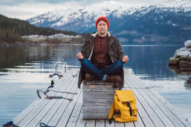 Gezgin adam, bir dağ ve gölün arkasındaki ahşap iskelede meditasyon pozisyonunda oturuyor. Metin iletiniz ya da tanıtım içeriğiniz için boşluk.