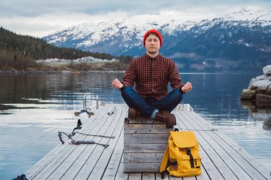 Gezgin adam, bir dağ ve gölün arkasındaki ahşap iskelede meditasyon pozisyonunda oturuyor. Metin iletiniz ya da tanıtım içeriğiniz için boşluk.