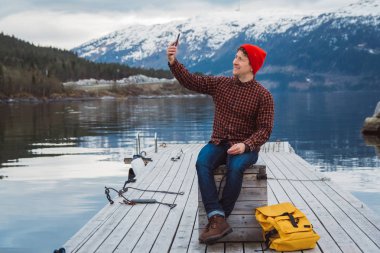 Gezgin adam akıllı telefonla kendi portresini çekiyor. Sarı bir sırt çantası bir dağ ve bir göl arka plan ahşap iskele üzerinde oturan Turist. Gezgin yürür, dağlarda fotoğraf çeker.