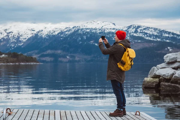 Traveler man taking photo with smartphone on mountains. Tourist in yellow backpack standing on a background of a mountain and a lake a wooden pier. Traveler walks, takes photo in mountains. View from