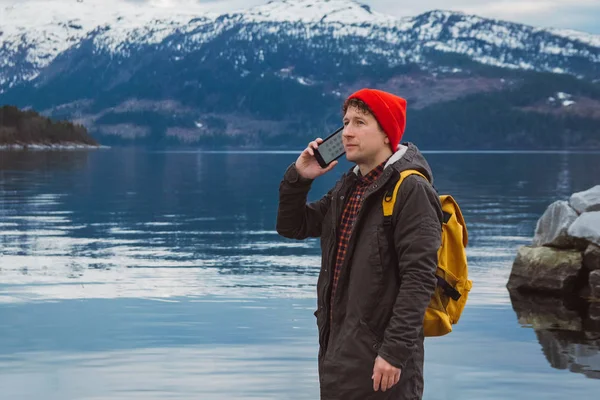 Traveler man talking on mobile phone. Tourist in a yellow backpack standing on a background of a mountain and a lake. Traveler walks, smiling man calling friends. Telephone conversation.