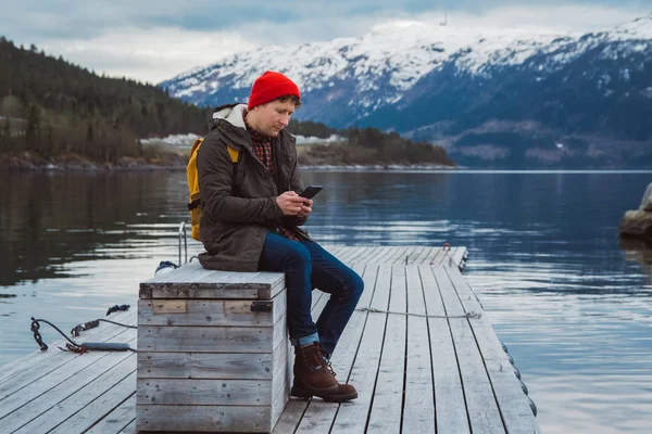 Traveler man looking into the phone. Tourist in yellow backpack sitting on a background of a mountain and a lake a wooden pier. Traveler walks, takes photo in mountains.